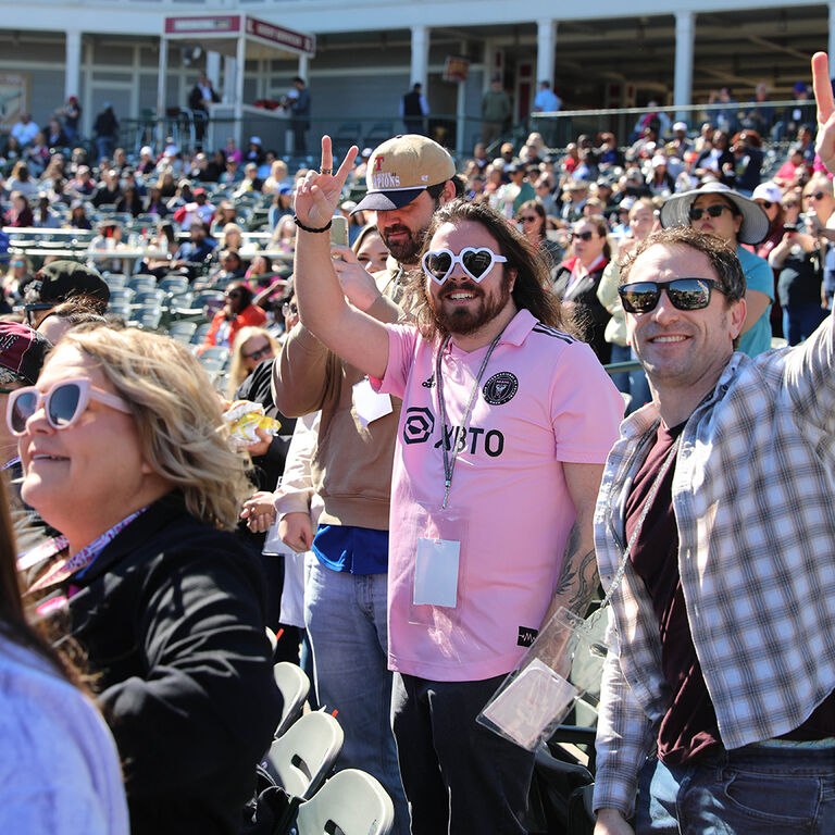 Crowd of people standing in stadium seating during an outdoor event, one person raising a peace sign